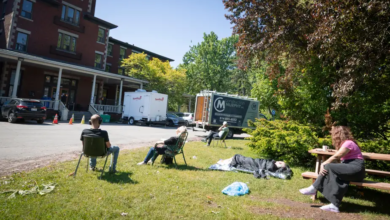 People sit on folding chairs and a bench in a grassy area outside a brick building, with trucks in the background and one person lying under a blanket on the ground near trees.