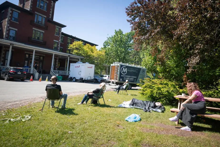People sit on folding chairs and a bench in a grassy area outside a brick building, with trucks in the background and one person lying under a blanket on the ground near trees.