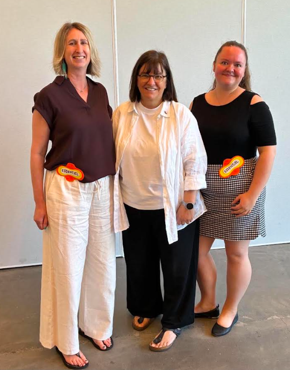 image Média Laval Three women smiling and standing together indoors; the two on the ends wear casual outfits with orange badges on their waists labeled 'ESSENTIEL'.