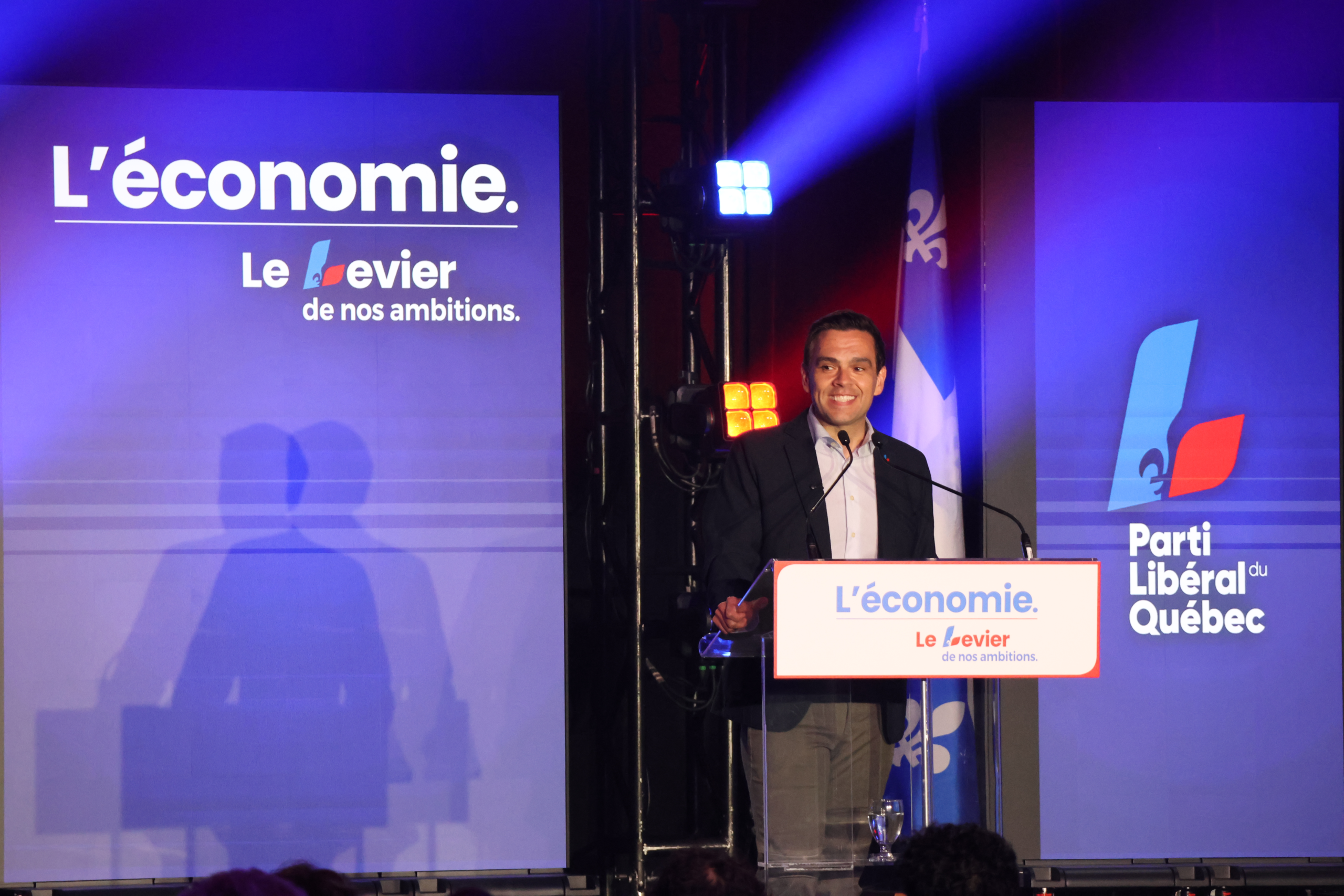 Man at a podium giving a speech on a blue campaign stage with 'L'économie' slogan and Parti Libéral du Québec banners