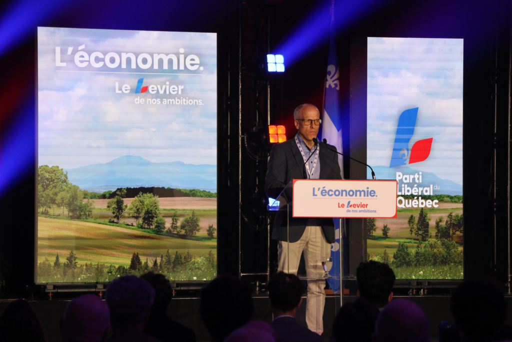Pierre Cossette candidat libéral dans Sherbrooke Média Laval Male speaker in a suit stands at a podium on a stage, with banners reading “L’économie” and the Quebec Liberal Party logo in the background.
