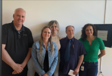 Five adults smiling and posing together in a bright office hallway.
