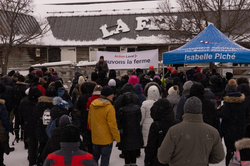 image Média Laval Crowd of people in winter coats gathered outdoors for a protest, facing a speaker; banner reads'Action Laval Sauvons la ferme' in front of a farm-like building, with a blue tent labeled 'Isabelle Piché' on the right.