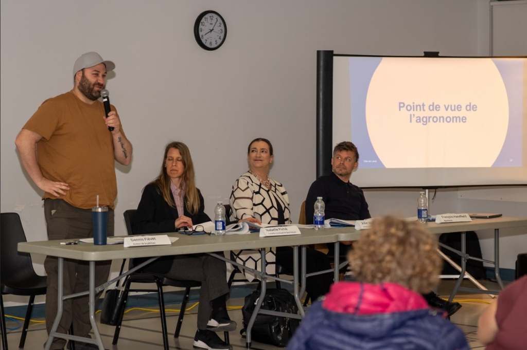 image Média Laval Panel discussion: man in brown T-shirt and cap speaks into a microphone at left while three panelists listen at a long table; projection screen behind them.