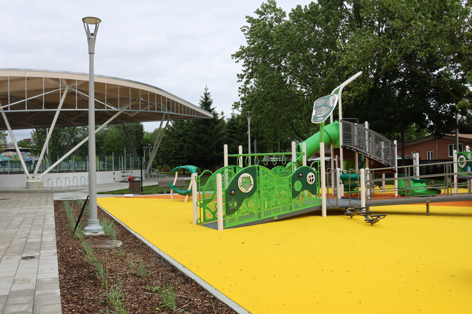 Children's playground with green climbing structure on a bright yellow safety surface near a covered seating area. Gently framed by trees and lampposts.
