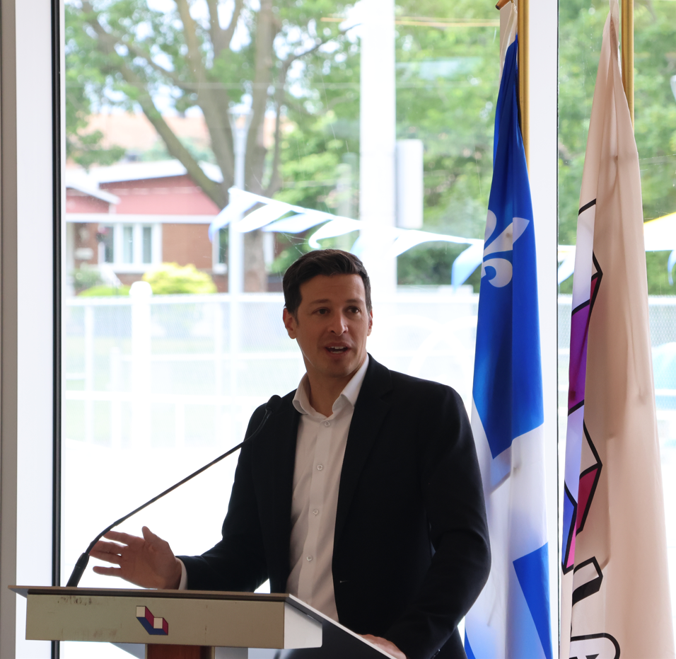 Man in a dark suit speaks at a podium with a microphone, gesturing with one hand during a formal event, with flags in the background.