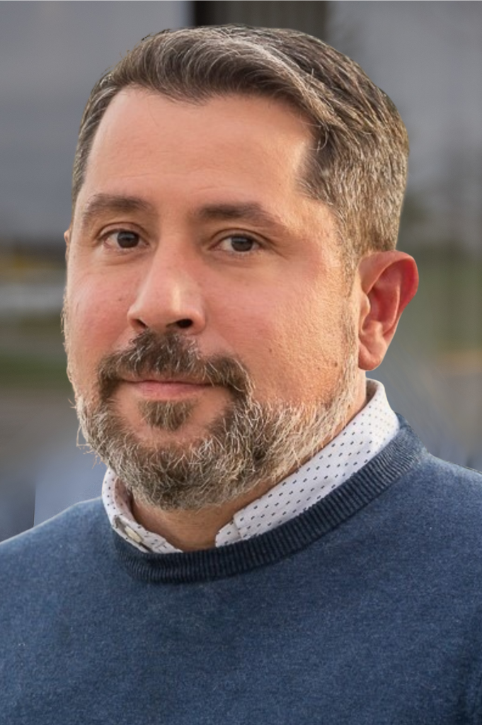 Portrait of a middle-aged man with short dark hair and a gray beard, wearing a blue sweater over a white dotted shirt, outdoors.