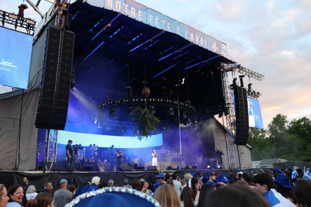 Outdoor concert stage with massive vertical speaker arrays, blue stage lighting, and a crowd watching the performance at dusk.