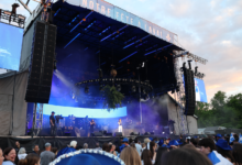 Outdoor concert stage with massive vertical speaker arrays, blue stage lighting, and a crowd watching the performance at dusk.