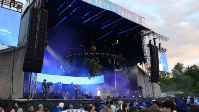 Outdoor concert stage with massive vertical speaker arrays, blue stage lighting, and a crowd watching the performance at dusk.