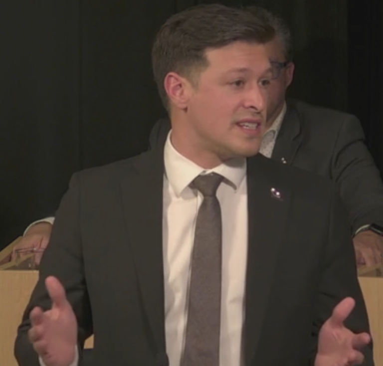 image Média Laval Man in a dark suit and tie speaking on stage, gesturing with his hands while addressing an audience.