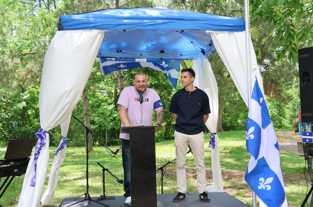 Two men on an outdoor stage under a blue canopy, one speaking into a microphone at a black lectern while the other watches nearby.