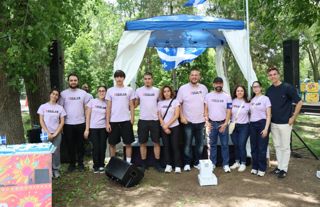 Twelve volunteers in lavender SCALAR shirts pose together outdoors under a blue canopy at a park event, with a colorful cake table to the left.