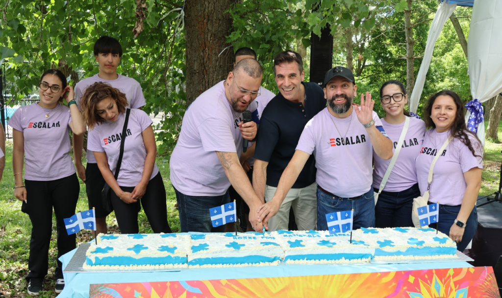 Eight people in lavender Scalar shirts join hands to cut a large blue-and-white frosted cake with blue flags at an outdoor park.