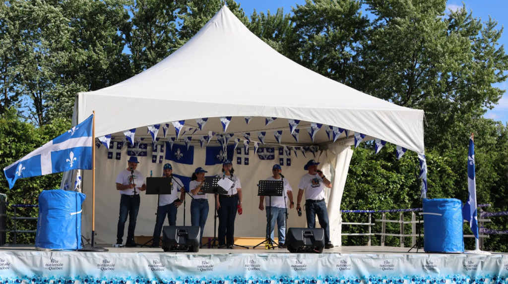 Seven-person band performing on an outdoor stage under a white canopy, with blue-and-white bunting and Quebec flags in the background.