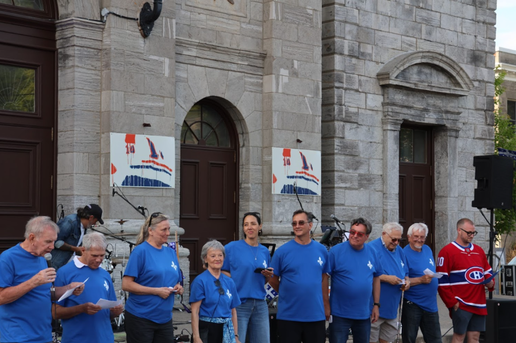 Group of people wearing matching blue shirts on a stage outside a stone building, some holding papers, preparing to perform.