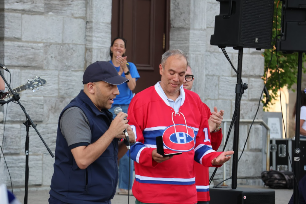 Man in a navy vest and cap speaks into a microphone at an outdoor event, standing next to a man in a red hockey jersey who holds a small trophy or award.
