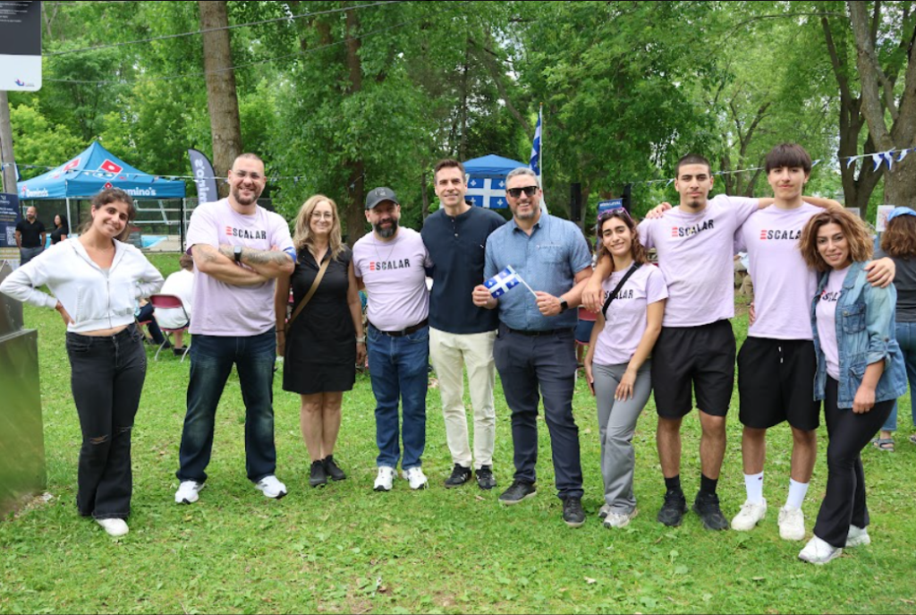 Ten people stand together in a park with tents and trees behind them, smiling for a group photo; several wear light pink shirts with a logo, one holds a Greek flag.