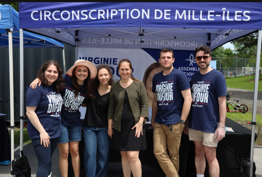 Six volunteers posing under a blue canopy reading Circonscription de Mille-Îles, wearing navy shirts and smiling outdoors at a community event.