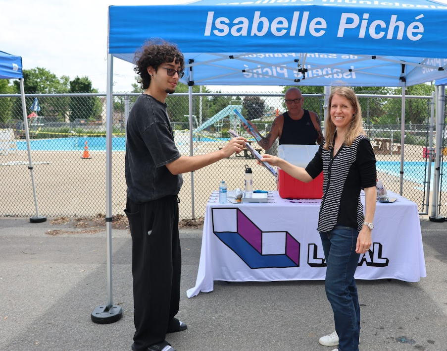 Man in black clothing hands a flyer to a smiling woman at an outdoor event booth under a blue canopy reading Isabelle Piché on top edge.