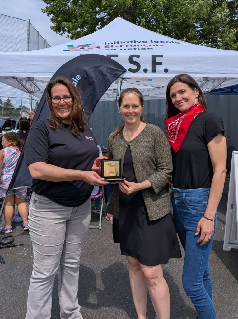 Three women pose at an outdoor event under a white canopy; one hands a small award to another, while a third smiles nearby.