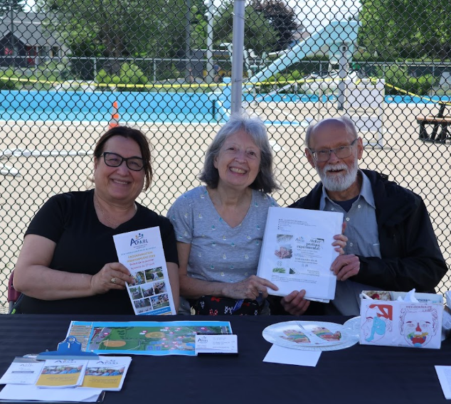 Three adults sit at a table outdoors by a chain-link fence and pool; left woman with glasses holds a brochure, center woman smiles, man on the right holds papers.