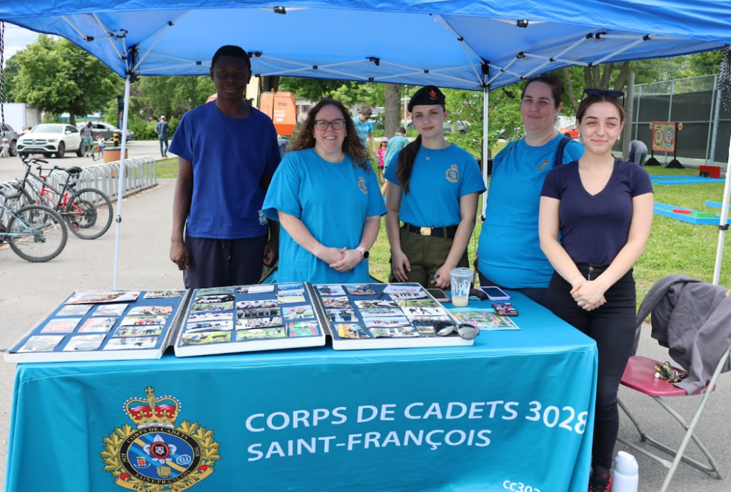 Five cadets stand behind a blue table under a canopy, displaying photo albums at a community event. Banner reads'Corps de Cadets 3028 Saint-François.'