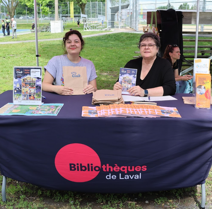 Two women sit at a blue-clothed table with library pamphlets and bags at an outdoor Bibliothèques de Laval booth in a park sunlit setting.