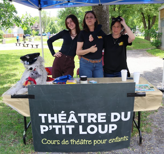 Three women at an outdoor booth for Théâtre du P'Tit Loup, with a plush wolf puppet on the table and a banner reading 'Cours de théâtre pour enfants'.