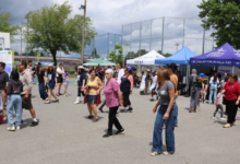 Crowd at an outdoor community fair with blue and white tents and people walking around on a sunny day.