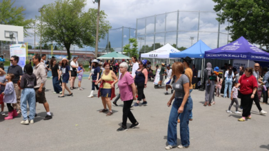 Crowd at an outdoor community fair with blue and white tents and people walking around on a sunny day.