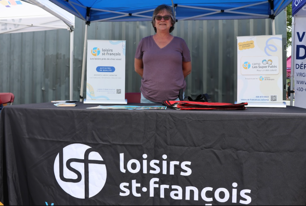 Woman in a purple T-shirt and sunglasses stands behind a black table with Loisirs St-François branding under a blue canopy at an outdoor information booth.