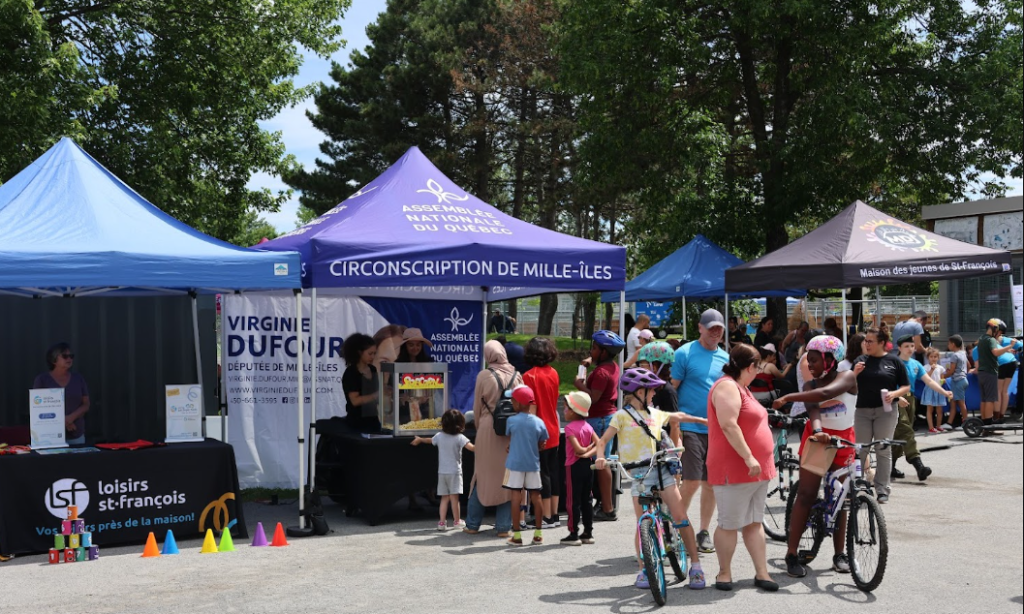 Community fair with colorful tents and families gathered, children lining up at a stall under a purple tent labeled Circonscription de Mille-Îles.