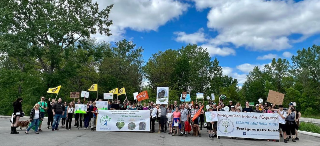 Coalition Mobilisations Citoyennes Environnementales de Laval Média Laval Group of diverse people holding environmental protest banners and signs outdoors on a sunny day with trees in the background.
