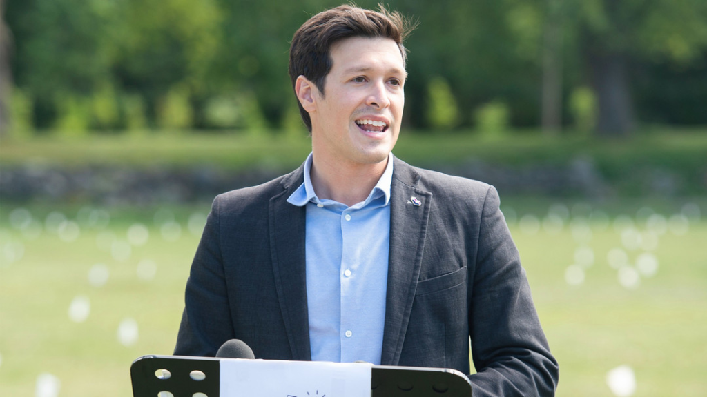 Man in a blazer speaks outdoors with a microphone, in a park setting behind him