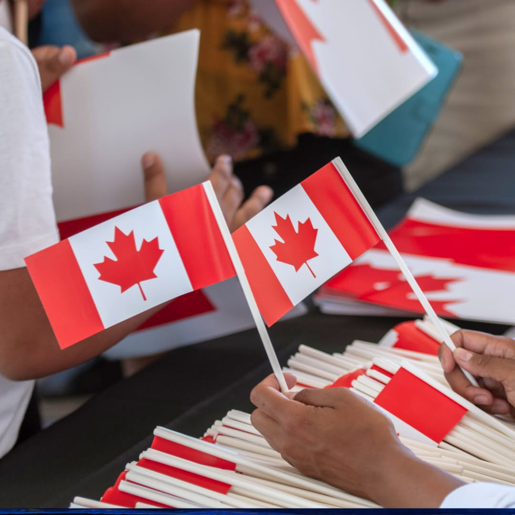 People hand out small Canadian flags on sticks at a table, with flags and stacks visible in the foreground.