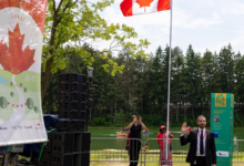 Man in a suit waves at the camera during an outdoor event by a lake, with a Canadian flag, large speaker stack and festival banners nearby.