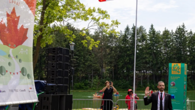 Man in a suit waves at the camera during an outdoor event by a lake, with a Canadian flag, large speaker stack and festival banners nearby.