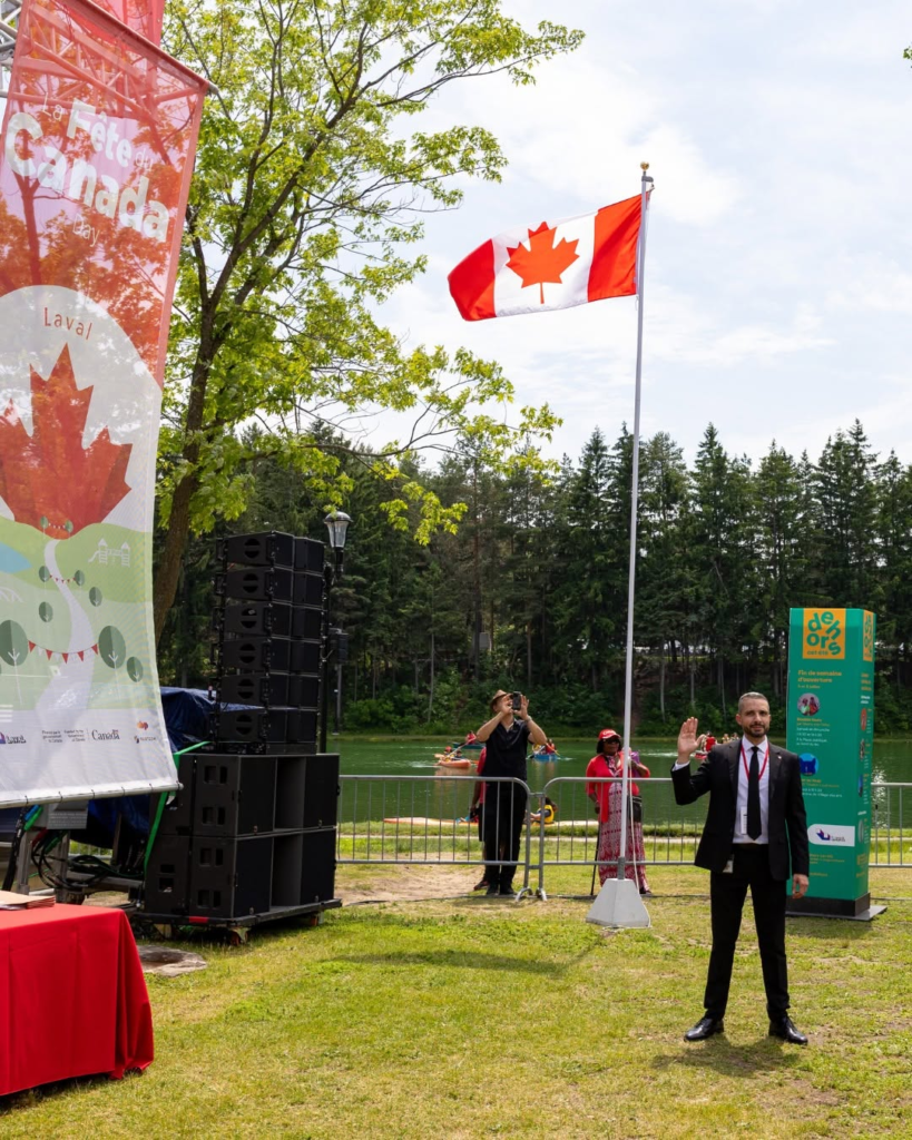 Man in a suit waves at the camera during an outdoor event by a lake, with a Canadian flag, large speaker stack and festival banners nearby.