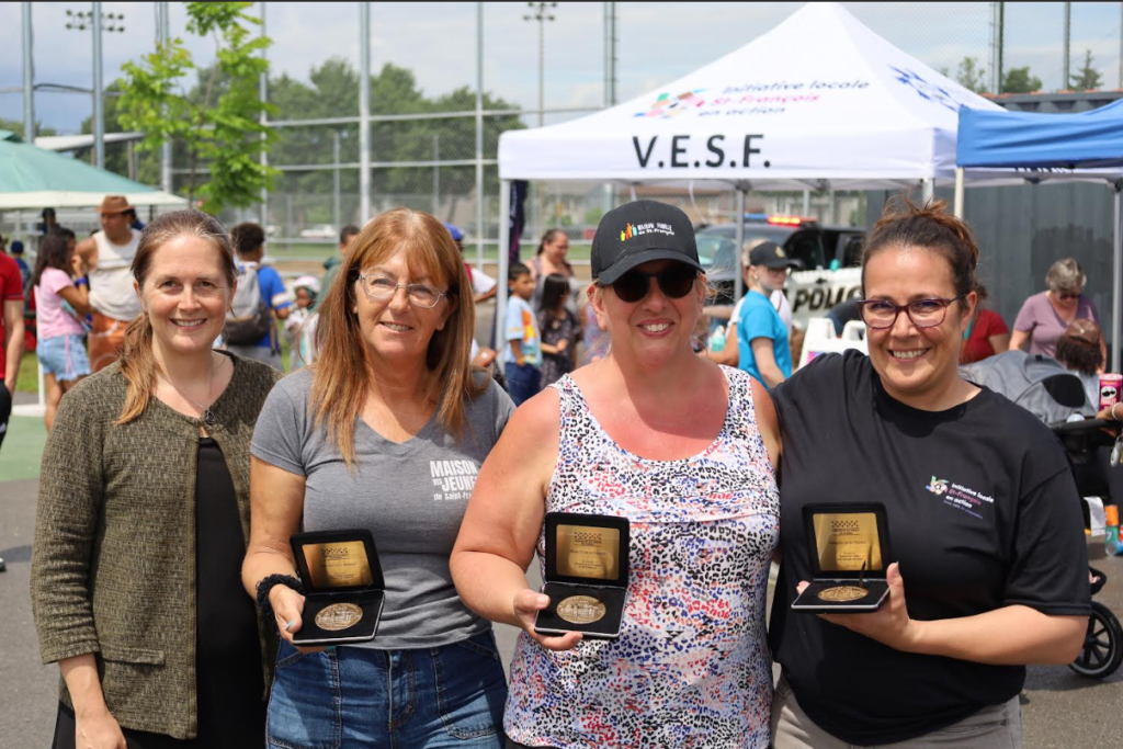Four smiling women pose with medals in boxes at an outdoor community event, with a white V.E.S.F. tent in the background.