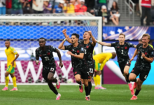 Five soccer players in black uniforms celebrate a goal, sprinting past the goal with raised arms and fans in the background.