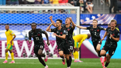 Five soccer players in black uniforms celebrate a goal, sprinting past the goal with raised arms and fans in the background.