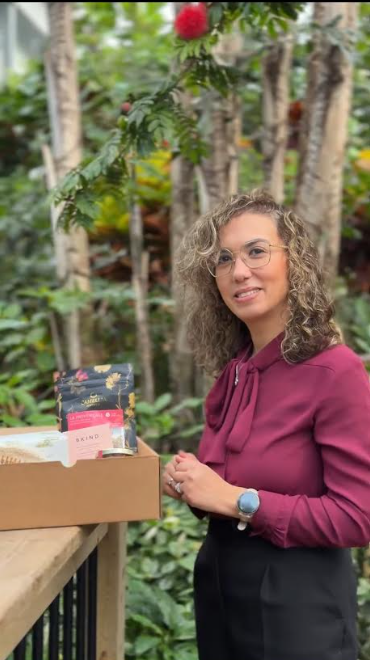 Woman with curly hair and glasses, wearing a magenta blouse, stands beside an open box of items on a wooden railing in a lush garden.
