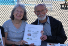 Two smiling seniors sit at a table outdoors behind a chain-link fence, holding a document; a pool and slide are visible in the background.