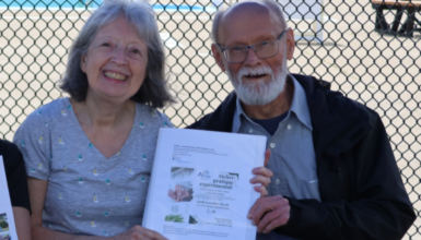 Two smiling seniors sit at a table outdoors behind a chain-link fence, holding a document; a pool and slide are visible in the background.