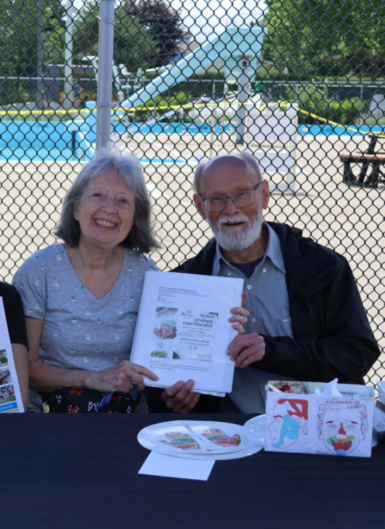 Two smiling seniors sit at a table outdoors behind a chain-link fence, holding a document; a pool and slide are visible in the background.
