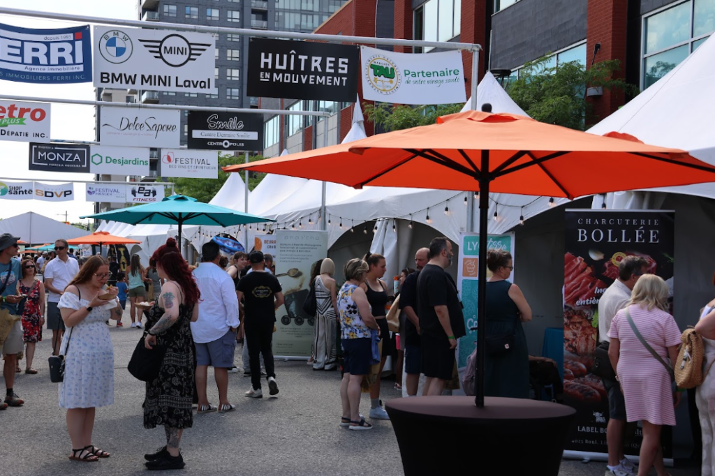 Crowd browsing stalls at an outdoor street market with white tents and orange and teal umbrellas in the foreground.