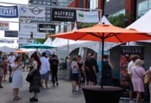 Crowd browsing stalls at an outdoor street market with white tents and orange and teal umbrellas in the foreground.