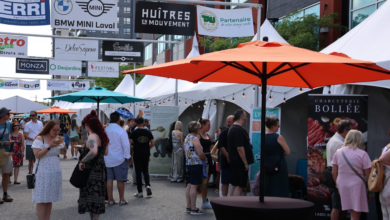 Crowd browsing stalls at an outdoor street market with white tents and orange and teal umbrellas in the foreground.