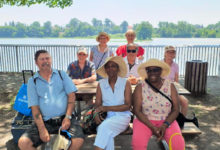 Group of seniors posing at a lakeside park picnic table under leafy trees, smiling with a sunny day by the water.